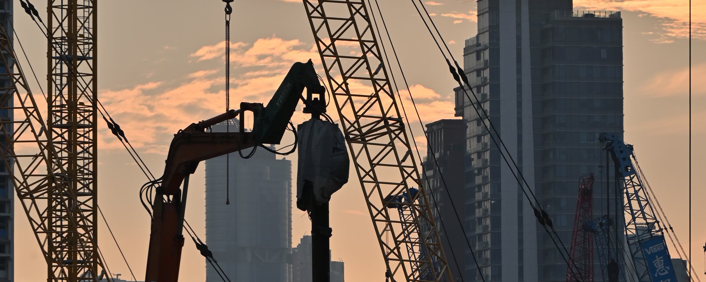 A city construction site at dusk: tower cranes, an excavator and high-rises against an orange sky